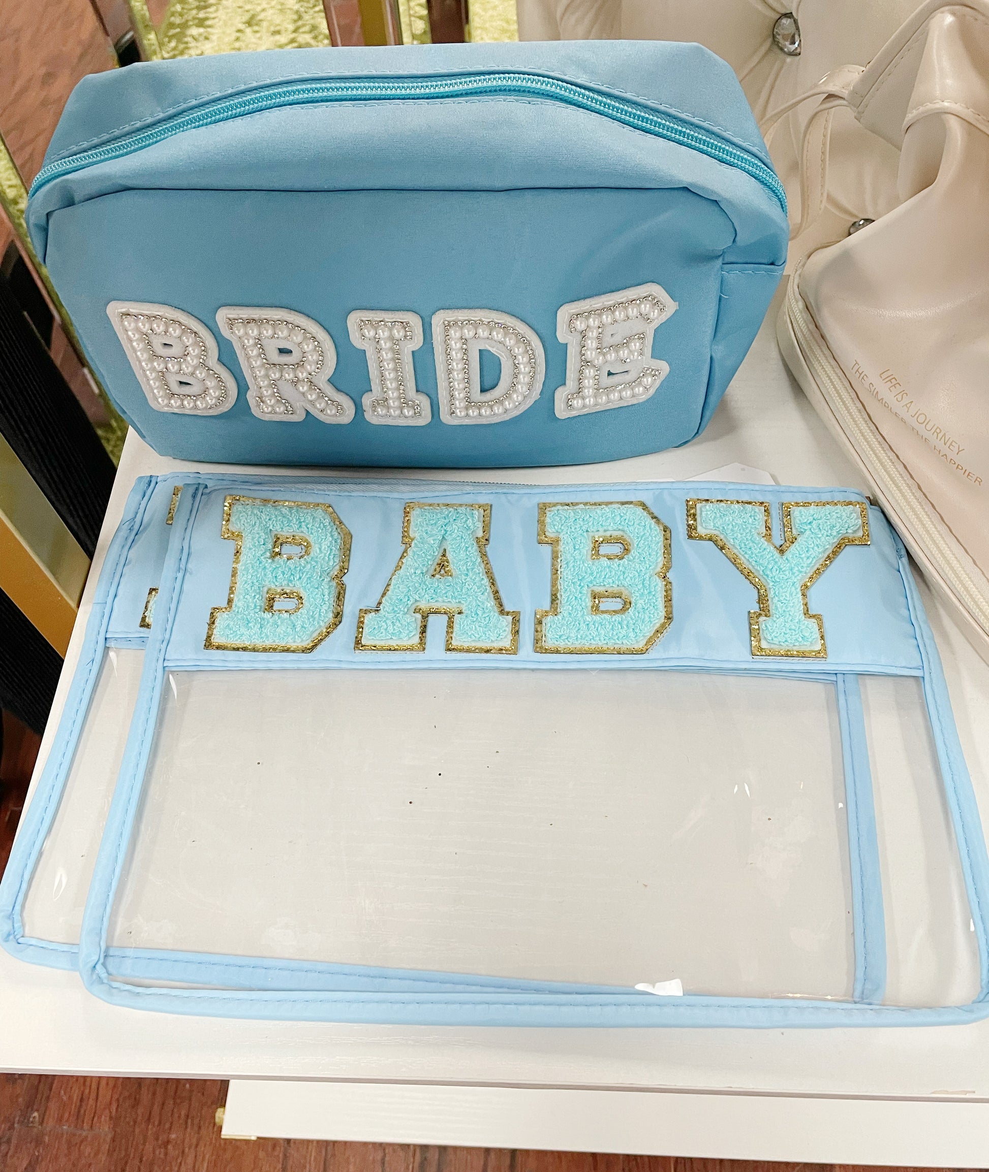 Clear zippered pouch that has a light blue border with the word 'baby' embroidered at the top in chenille light blue varsity letters outlined in shiny gold.